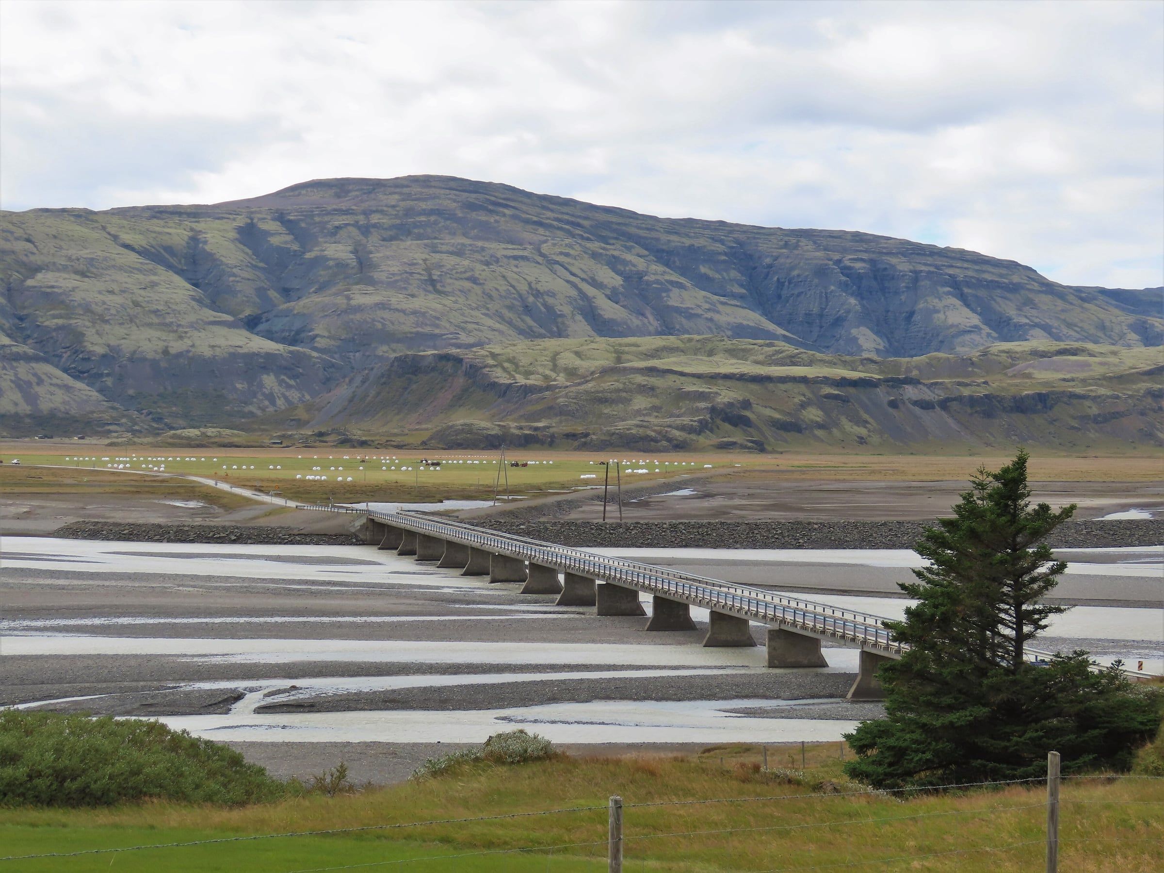 A long concrete road bridge crosses braided glacial river channels on a flat plain beneath a broad mountain ridge.