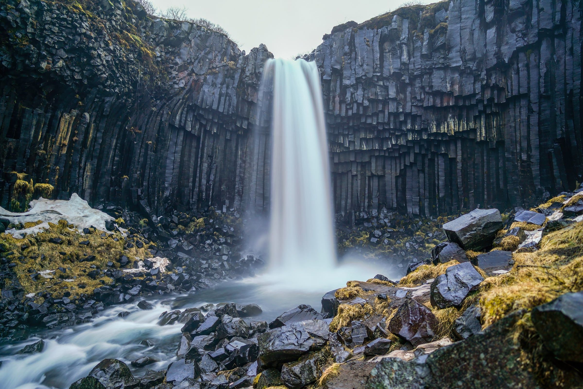 A white waterfall pours between tall dark basalt columns that rise above a rocky stream bed and green moss.